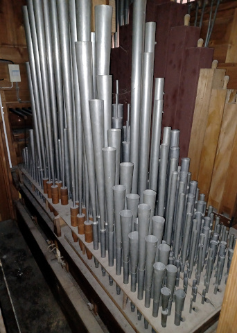 Une vue de la tuyauterie.
Celle-ci est disposée diatoniquement (alternativement d'un côté et de l'autre),
et en mitre (avec les basses au centre).
Le fond de l'orgue est à gauche de l'image, l'avant à droite. On distingue le revers
de la façade en haut, derrière les tuyaux rouges du Bourdon.
Tout à gauche, le revers de la console, avec la mécanique de tirage des jeux.
Au premier plan, la Trompette, dont les 12 tuyaux graves, avec leurs pieds en bois tournés,
sont vraiment caractéristiques de la facture des Callinet.
La tuyauterie a visiblement subi des accords trop vigoureux (déchirures),
mais ne semble pas avoir été altérée ou subi de changement de diapason.