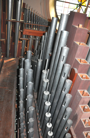 Le sommier des "petits jeux" du grand-orgue
est parallèle au précédent (dont on aperçoit l'extrémité tout à gauche,
au bout de la passerelle qui les sépare).
La vue est tournée de 90° par rapport à la précédente, mais au même endroit.
On tourne donc le dos à la façade.
En haut, le fond de la tribune, avec l'oculus et le Cornet
que l'on verra en gros plan sur la vue suivante.
Ce sont ici les basses des "petits jeux" (les tuyaux ne sont donc pas
si petits que cela). De gauche (passerelle) à droite (centre le l'orgue) :
la Doublette, le Nasard, la Flûte 4', le Principal 4',
et le Bourdon 8', en bois.